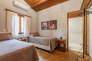 Une chambre avec deux lits, un plafond en bois, un climatiseur, une fenêtre avec des rideaux et deux tableaux encadrés au mur.