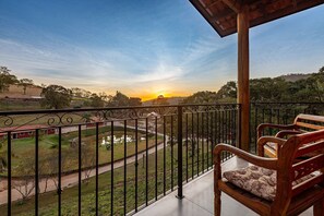 A balcony with a wooden chair, a view of a park with a pond, and a sunset in the background. - Fazenda Realeza (Pedra Bela)