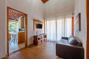A living room with a wooden floor, a brown sofa, a TV mounted on the wall, and a kitchen area visible through an open doorway. - Fazenda Realeza (Pedra Bela)