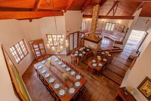 A spacious dining area with multiple tables set for a meal, a large stone fireplace, and a high ceiling with wooden beams. - Fazenda Realeza (Pedra Bela)