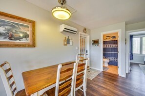 Un coin repas avec une table et des chaises en bois, un appareil de climatisation, une œuvre d’art encadrée et une salle de bain visible à travers une porte ouverte.