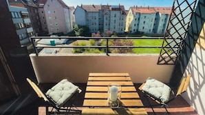 Un balcon avec une table en bois et deux chaises, un vase en verre contenant une plante, et une vue sur des immeubles et un espace vert.