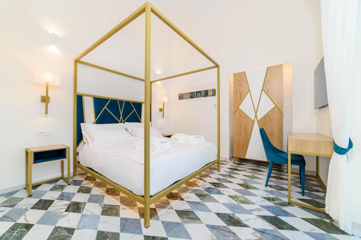 A room with a bed, bedside table, chair, desk, and artwork on the wall.