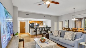 A modern living room with a grey sofa, a coffee table, and a TV. There is a kitchen in the background with a dining area. - Escape to Luxury: Spacious 3-Bedroom Condo, Perfect for Relaxation & Fun at the Sandestin Golf and Beach Resort! (Miramar Beach)