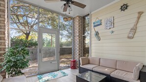 A sunlit room with a glass door leading to a backyard, a beige sofa, a glass coffee table, and nautical-themed wall decor.