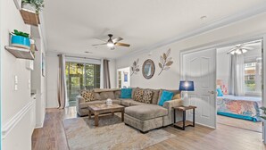 A living room with a sectional sofa, a coffee table, and a lamp. There is a ceiling fan and a door leading to another room.