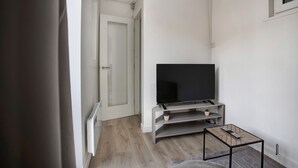 A modern living room with a flat-screen TV on a wooden stand, a small table with a remote, and a door leading to another room. - Cozy 1-bed Apartment Close to Lcy & Excel (London)