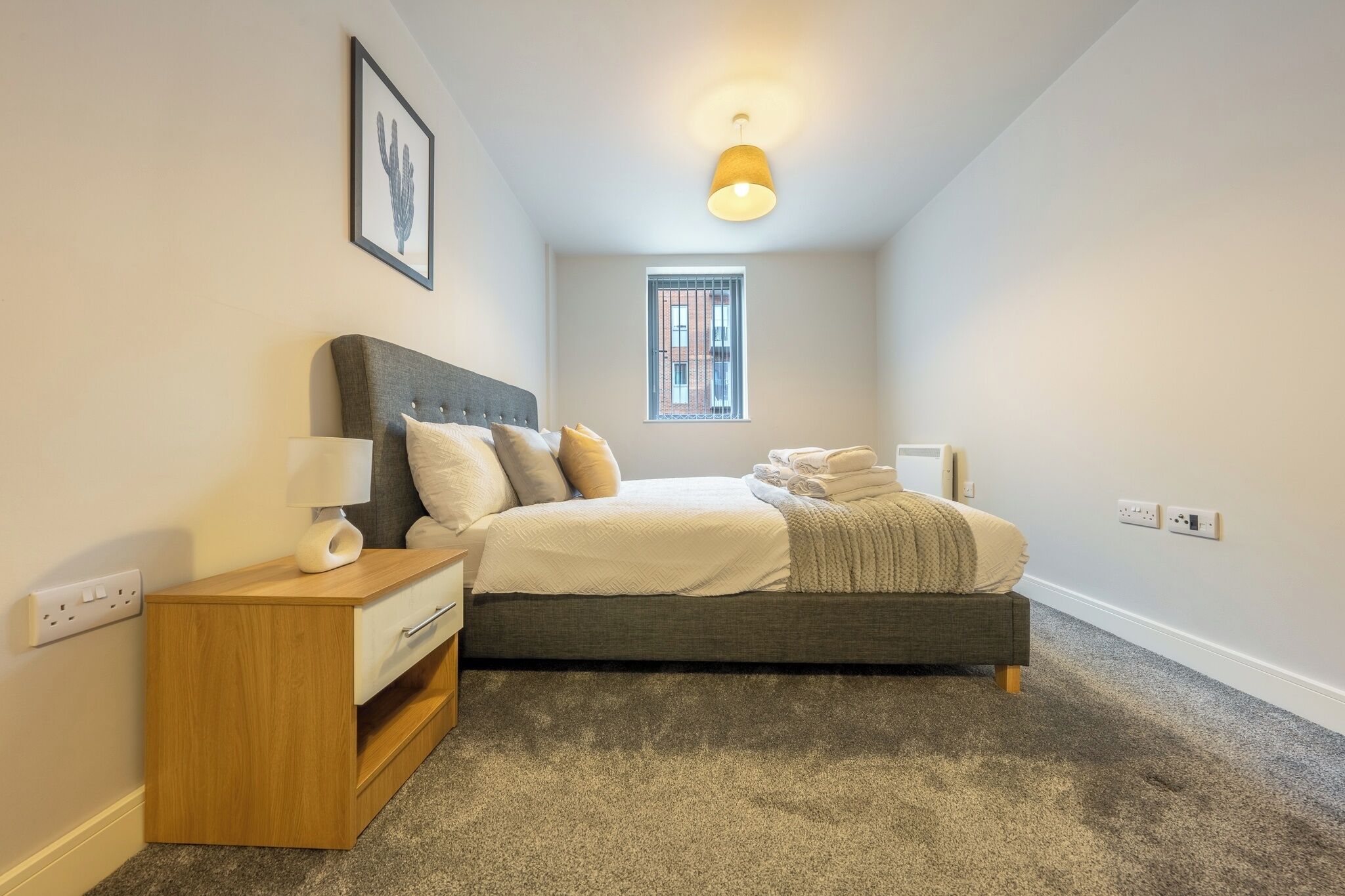 Une chambre à coucher avec un lit, une table de chevet, une lampe et une fenêtre donnant sur un immeuble.