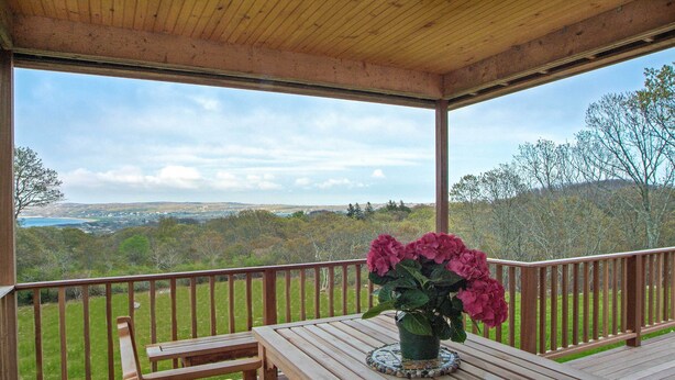 A wooden deck with a table and bench, a potted plant, and a view of a landscape with trees and hills. - Beautiful Aquinnah Home: Ocean View, Walk to Beach (Aquinnah)