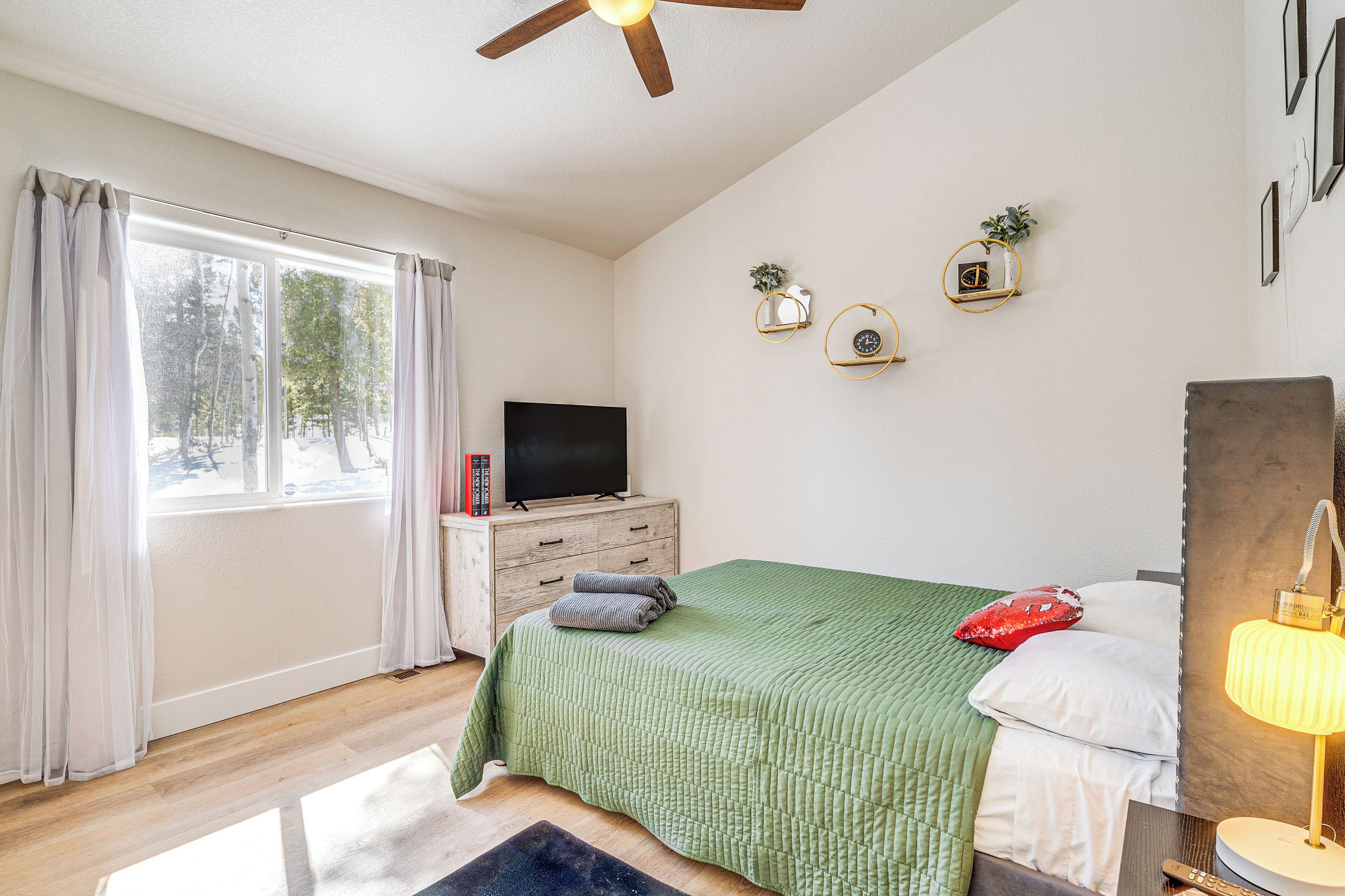 A bedroom with a green bedspread, a flat-screen TV on a wooden dresser, a bedside lamp, and a window with curtains.