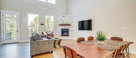 Amplia sala de estar con una gran mesa de comedor de madera, asientos cómodos, chimenea y ventanas grandes que ofrecen vistas a árboles.