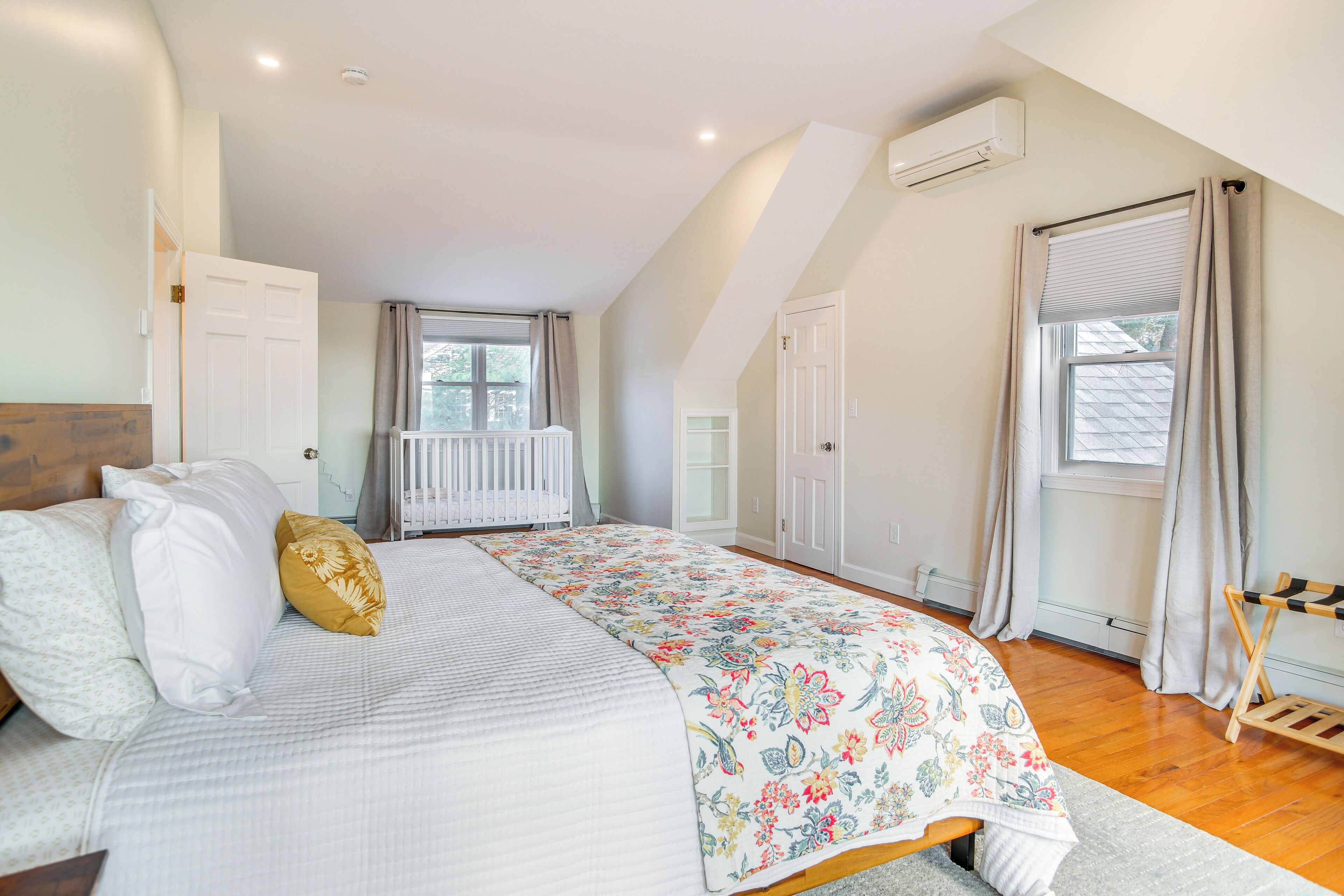 A bedroom with a large bed, a crib, a window with curtains, and an air conditioning unit on the wall.
