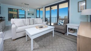 A living room with a white sofa, a coffee table, and a chair with a star-patterned pillow. There is a balcony with a view of the sky and a railing.
