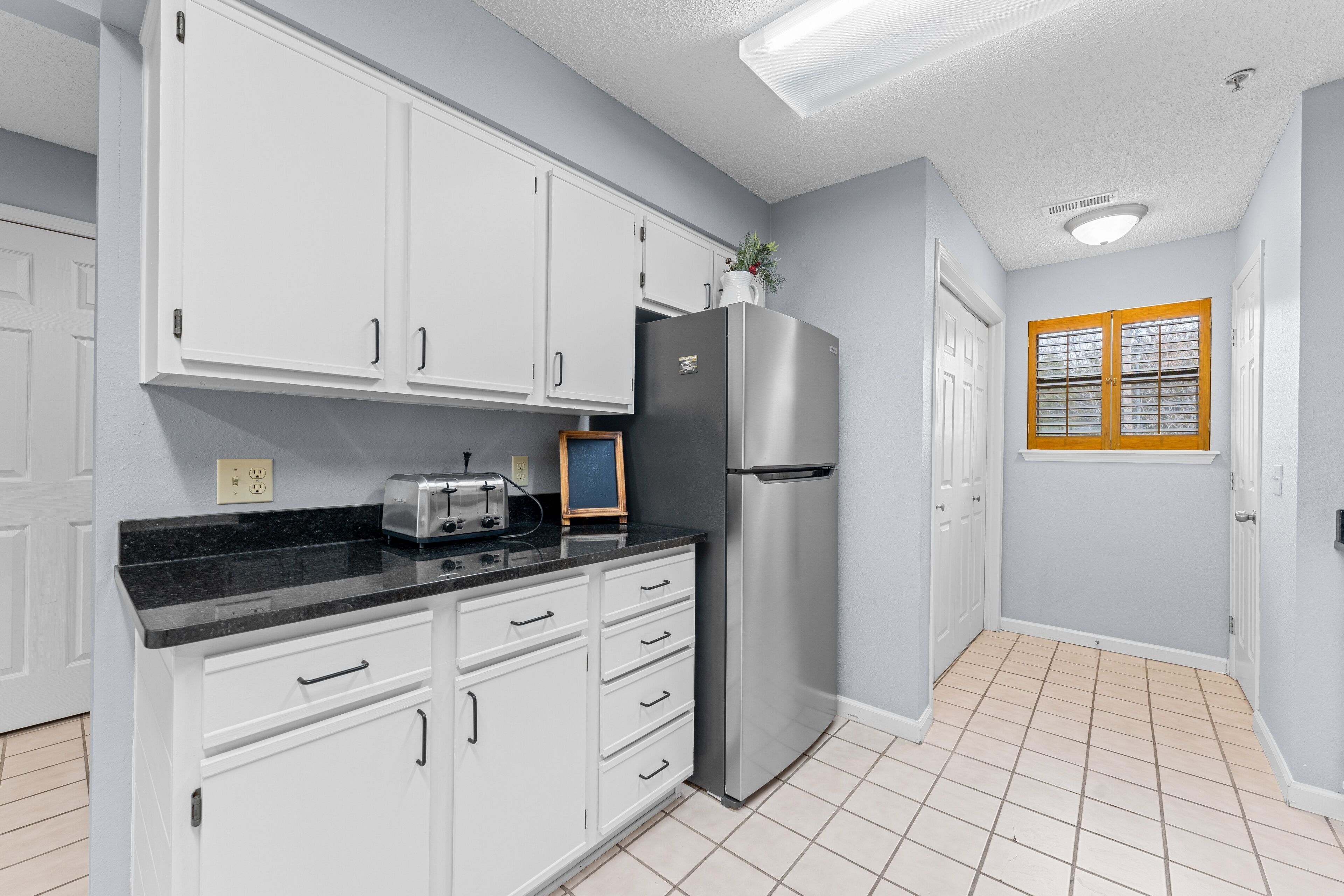 A kitchen with white cabinets, a stainless steel refrigerator, a black countertop, and a window with wooden shutters.