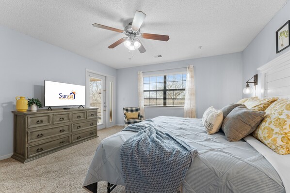 A bedroom with a bed, a ceiling fan, a dresser, a television, and a window with curtains.
