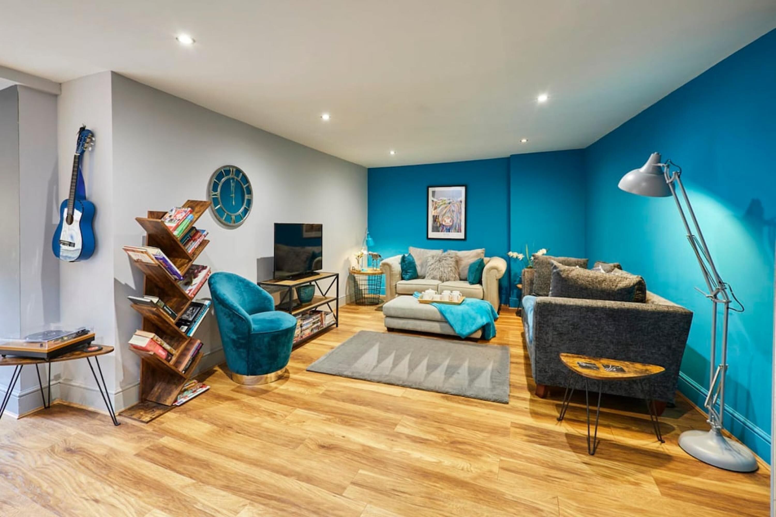 A modern living room with a blue accent wall, a guitar, a bookshelf, a sofa, a coffee table, and a floor lamp.