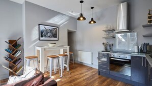A modern kitchen with a built-in oven, a coffee machine, and a stainless steel range hood. There are three stools around a white table, and a bookshelf with books to the left. - Host Stay Rooftops Grey (Alnwick)