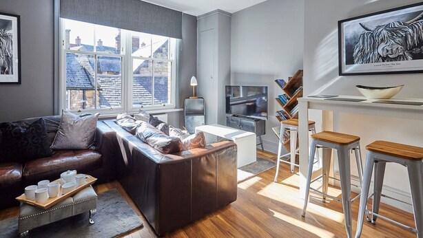 A modern living room with a leather sofa, a coffee table, and a TV. There is a window with a view of buildings outside. - Host Stay Rooftops Grey (Alnwick)