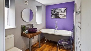 Un baño moderno con una bañera independiente, un espejo redondo, una pared de acento morada y un tocador de madera.