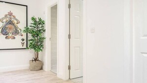 Un pasillo con una obra de arte enmarcada, una planta en maceta y una puerta blanca.