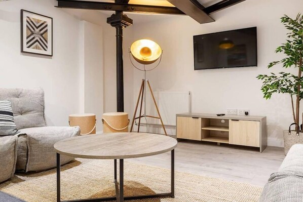 A modern living room with a round wooden table, two cushioned chairs, a floor lamp, a TV mounted on the wall, and a wooden cabinet.