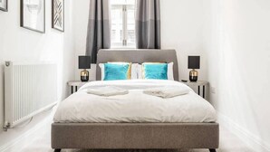 Un dormitorio moderno con una cama tapizada en gris, dos mesitas de noche con lámparas, un radiador y una ventana con cortinas grises.