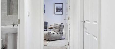Un pasillo que conduce a un baño con lavabo e inodoro, un sillón gris y una mesita.