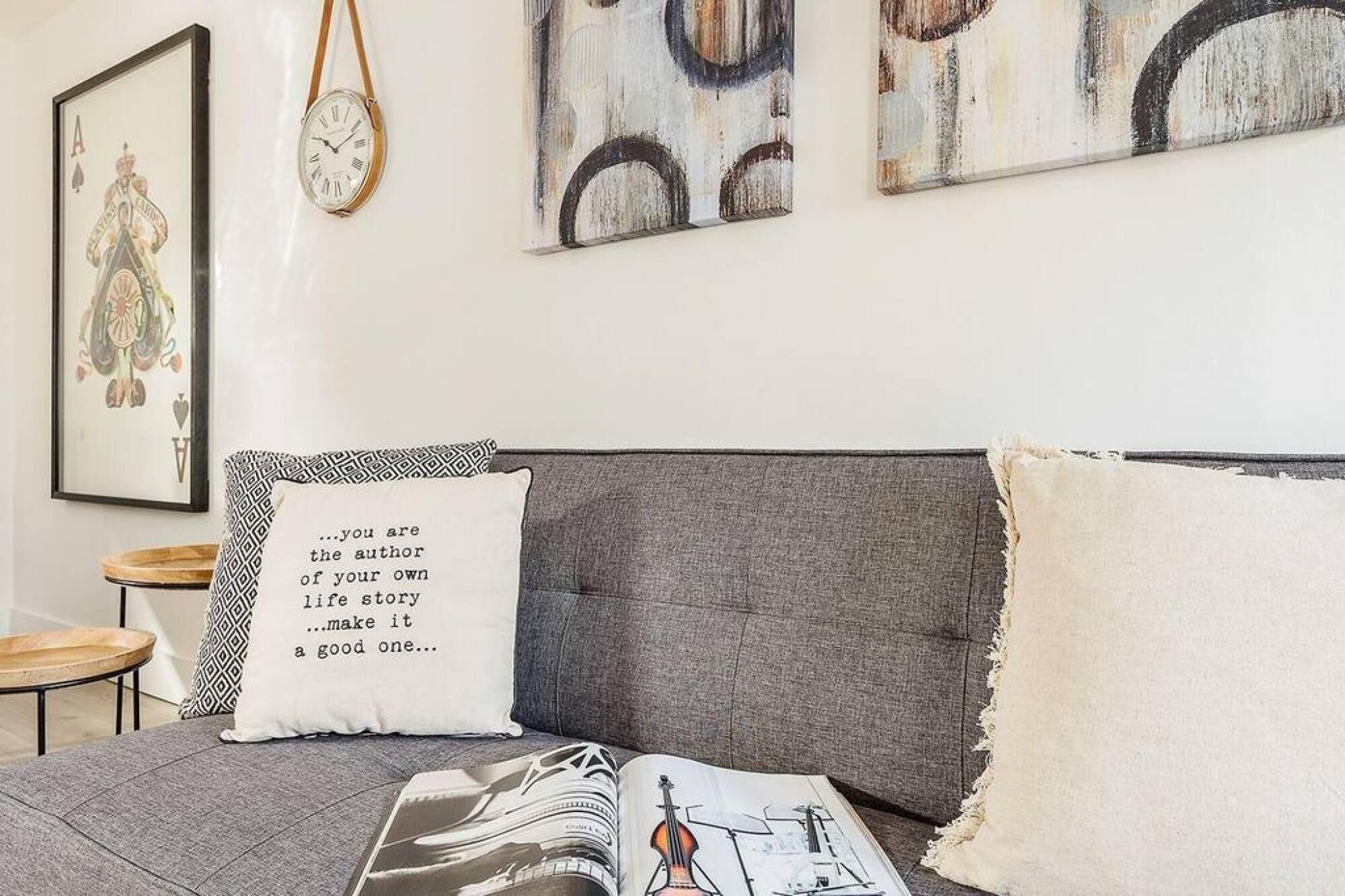 A bedroom with a grey headboard, a white pillow with a quote, and a framed playing card on the wall.