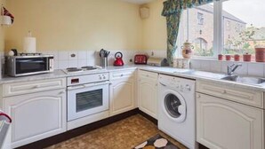 A compact kitchen with white cabinetry, a microwave, a toaster, a washing machine, and a window with a view of a building outside. - Host Stay Grange Cottage (Belford)