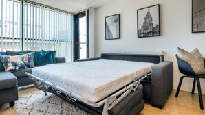 A modern bedroom with a convertible bed, a grey sofa, and a chair. There are two black-and-white framed pictures on the wall. - Host Apartments The Pall Mall Balcony Apt (Liverpool)