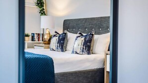 A modern bedroom with a grey upholstered bed, white linens, and two decorative pillows. A blue throw blanket is draped over the foot of the bed. A bedside table with a lamp and a small plant is visible. - Host Apartments The Pall Mall Balcony Apt (Liverpool)