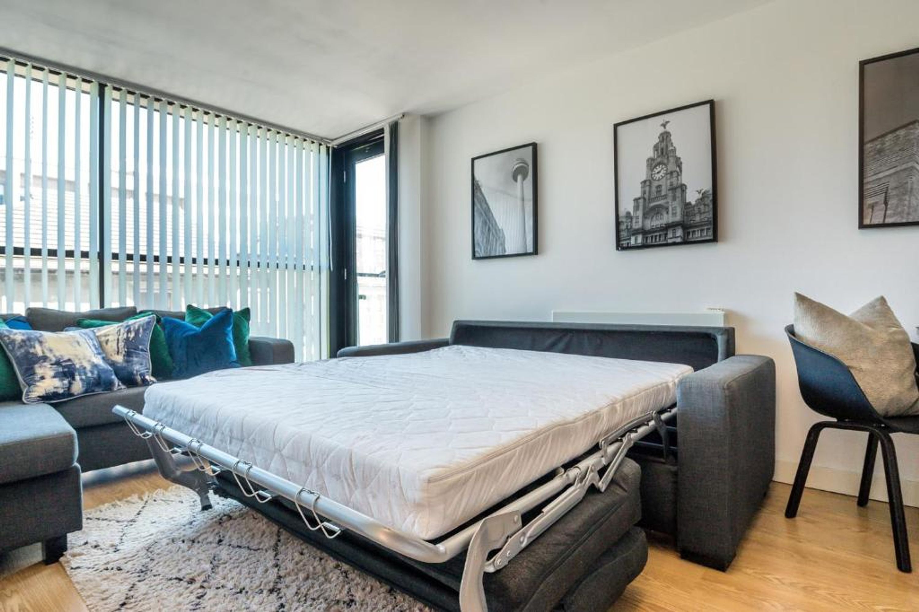 A modern bedroom with a convertible bed, a grey sofa, and a chair. There are framed pictures on the wall and a window with blinds.