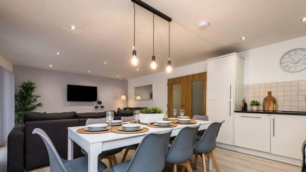 A modern kitchen with white cabinets, a dining table set for four, and a large clock on the wall. - Host Apartments Knight St Place (Liverpool)