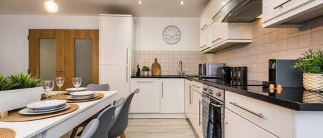 A modern kitchen with white cabinets, a black countertop, and a built-in oven. There is a dining table with chairs and a clock on the wall.