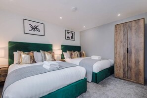 A hotel room with two beds, each with a green headboard, white bedding, and decorative pillows. There are two framed artworks on the wall, a wooden wardrobe, and a carpeted floor. - Host Apartments The Roscoe Coach House (Liverpool)