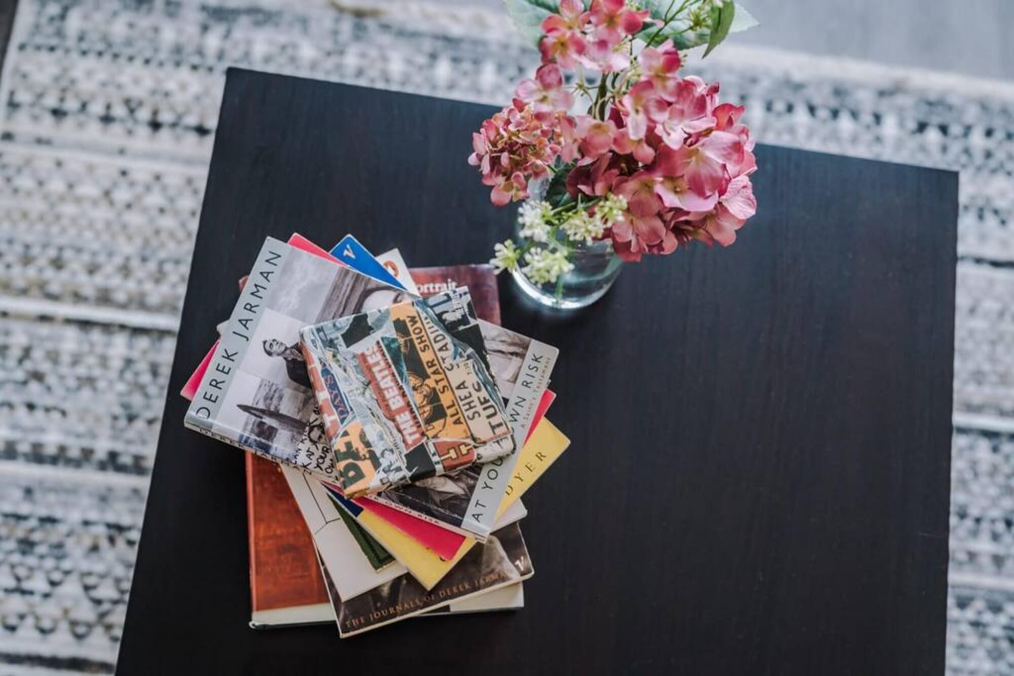 A stack of magazines and newspapers on a dark wooden table with a vase of pink flowers in the background.