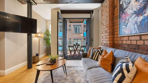 A modern living room with a sofa, coffee table, and a view of a brick building through an open door. - Host Apartments Queen Square Apartment (Liverpool)