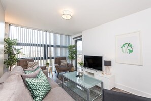 A modern living room with a sofa, armchairs, a glass coffee table, a flat-screen TV, and a large window with blinds.
