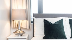 Cama bem arrumada com almofadas brancas e verde escuro, luminária de cabeceira e mesa de cabeceira branca.