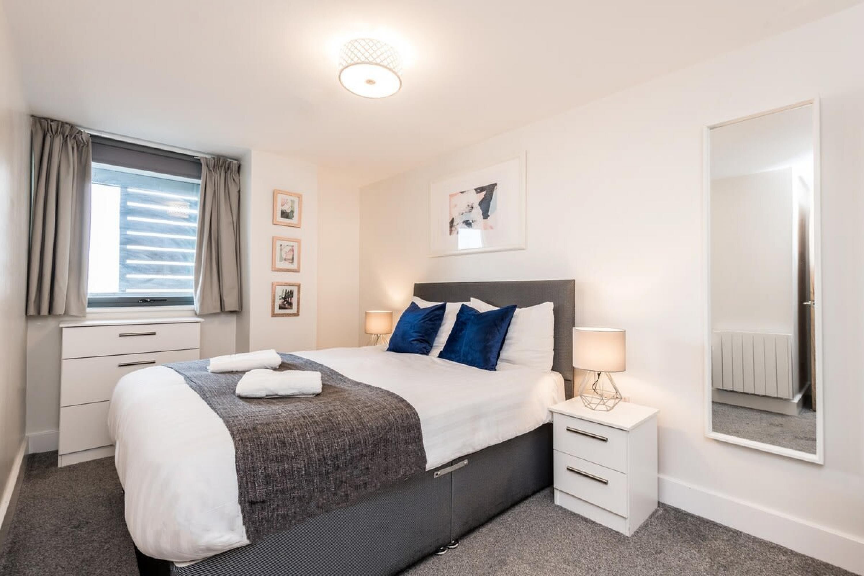 A modern hotel room with a large bed, two bedside tables, a window with curtains, and a mirror on the wall.