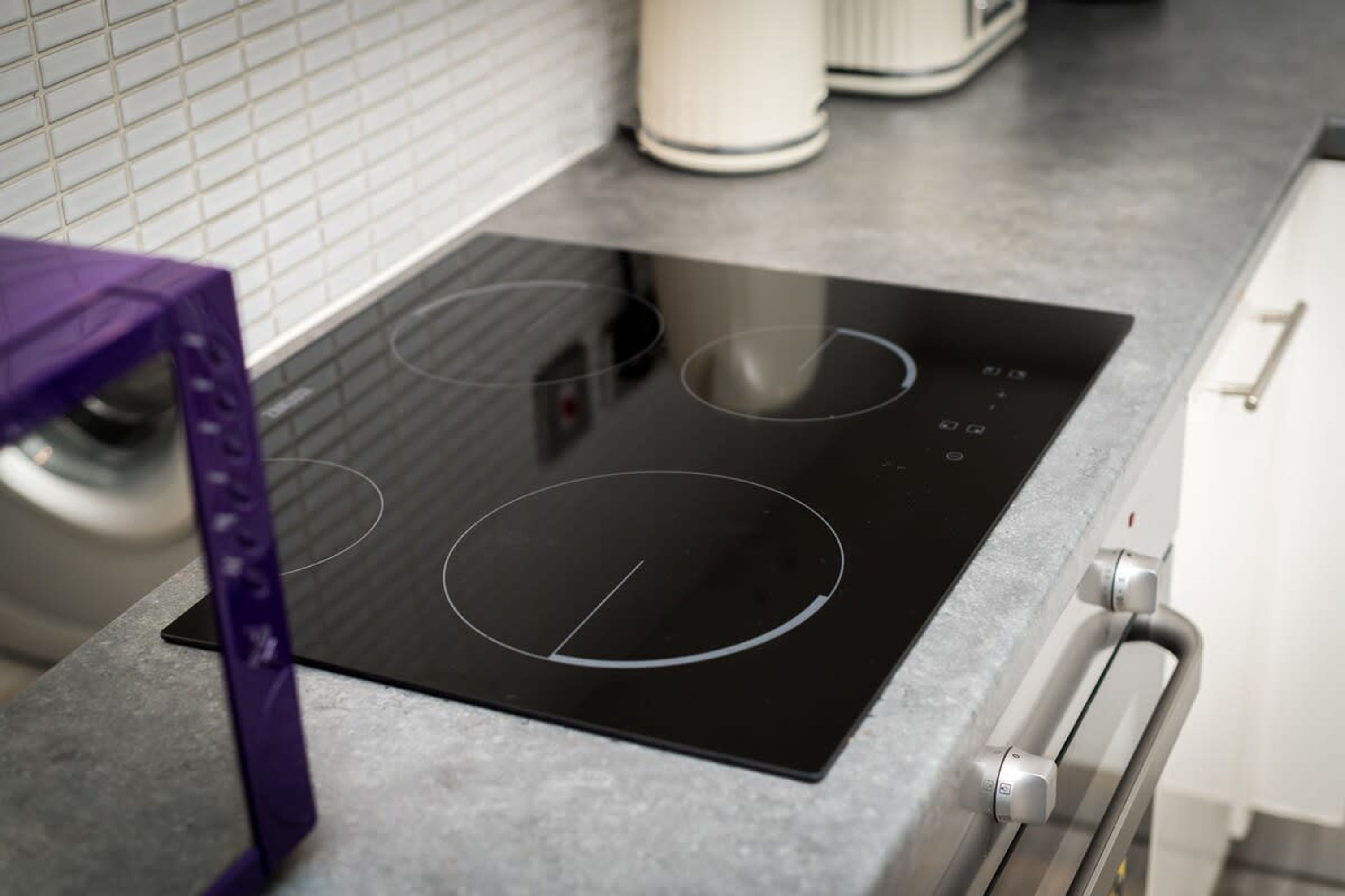 A kitchen with a black induction cooktop, a stainless steel oven, and a white tiled backsplash.