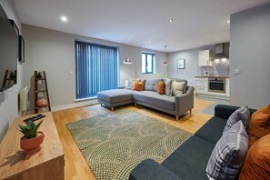En moderne stue med en hjørnesofa, et spisebord med en potteplante og et køkken i baggrunden.