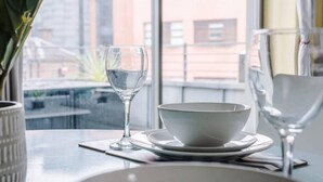 Une table à manger avec un bol et un verre d’eau, une plante en pot et une vue sur des bâtiments à travers une fenêtre.