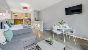 Un appartement moderne avec un canapé gris, un coin repas comprenant une table et des chaises blanches, une cuisine en arrière-plan et un téléviseur à écran plat fixé au mur.