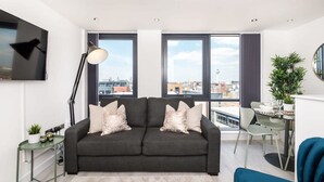 A modern living room with a dark grey sofa, a small side table, a floor lamp, and a dining area with a table and chairs. - Host Apartments The Baltic Penthouse 3 (Liverpool)