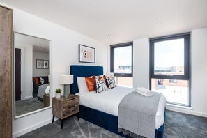 Quarto moderno com uma cama grande, mesa de cabeceira, espelho e duas janelas com vista para a cidade.