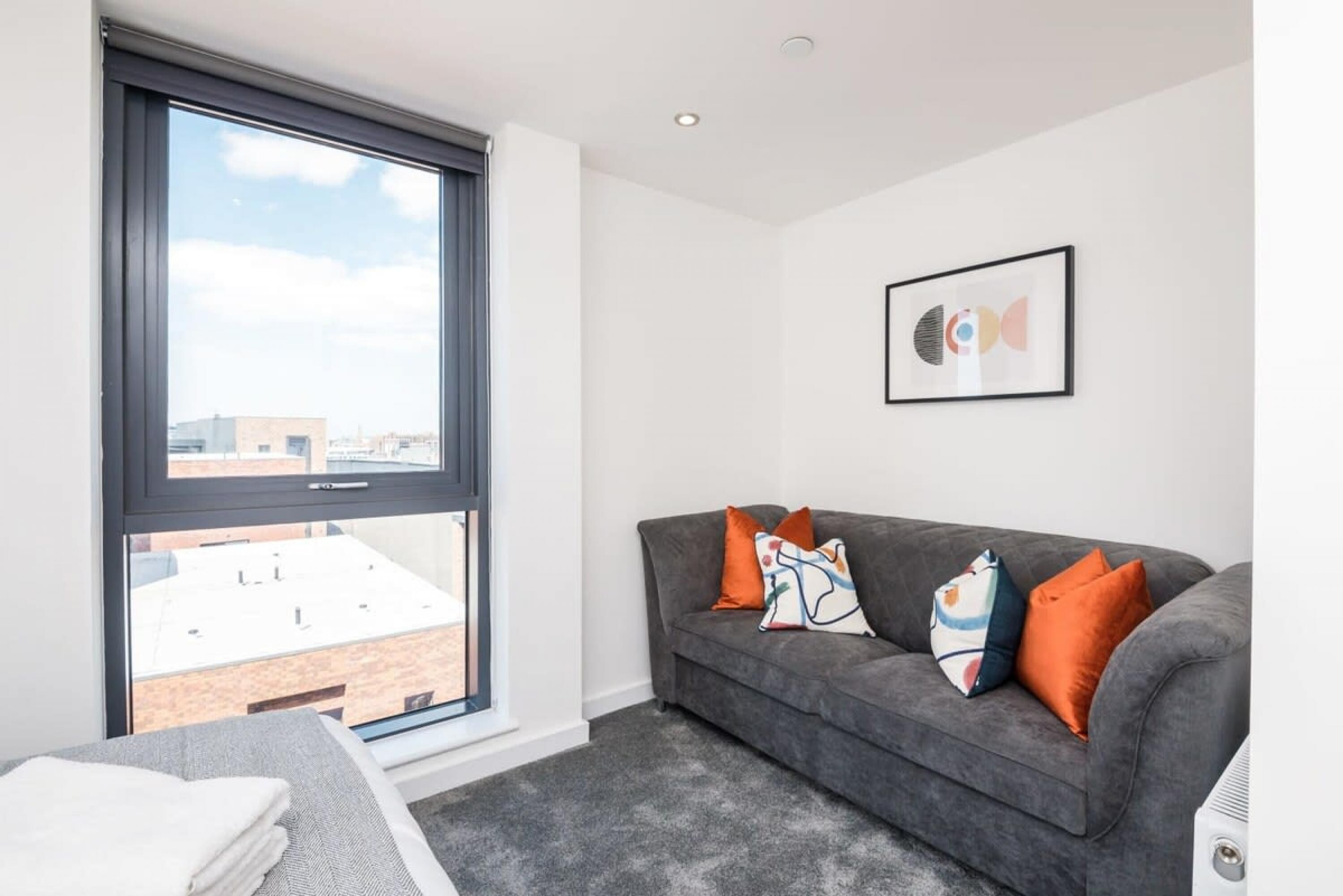 A modern living room with a grey sofa, colorful cushions, a framed abstract art piece, and a large window offering a city view.