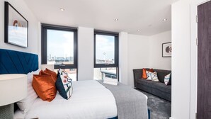 A modern hotel room with a large bed, a sofa, and two windows offering a city view. - Host Apartments The Baltic Penthouse 3 (Liverpool)