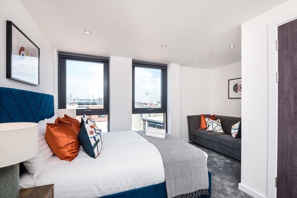 Quarto de hotel moderno com uma cama grande, um sofá e duas janelas com vista para a cidade.
