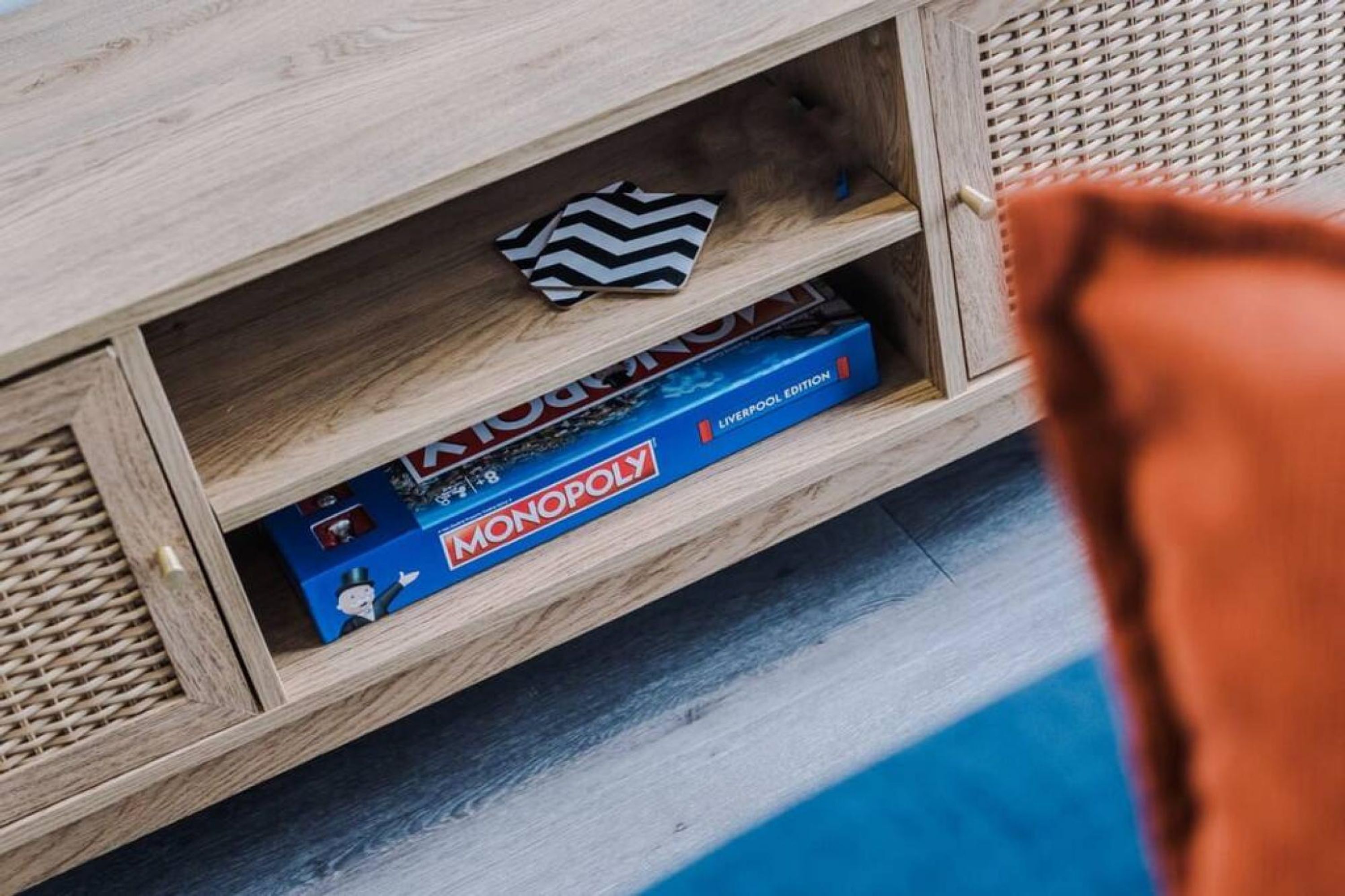 A wooden shelf with a board game, a striped coaster, and a book titled "Monopoly Liverpool Edition".